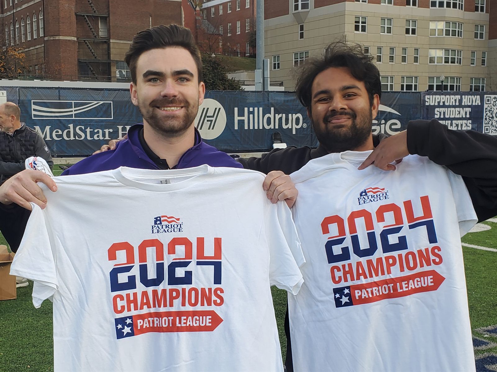 Dylan and a teammate on the field holding 2024 Patriot League Champions shirts after the title-clinching game.