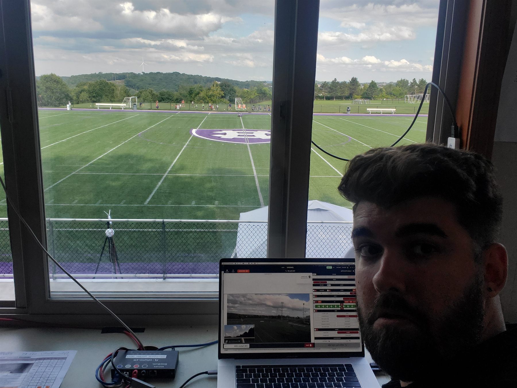 Dylan at the live-clipping station on the Holy Cross sideline, laptop open with the field beyond the booth window